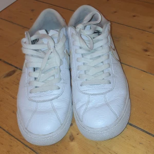 Vita Nike sneakers med gulddetaljer - Säljer ett par vita Nike sneakers med snygga gulddetaljer. Skorna har klassisk snörning och är perfekta för en stilren look. De är tillverkade i skinn och har en bekväm passform.