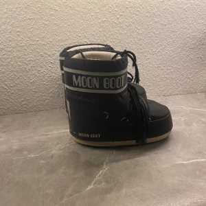 Svarta Moon Boots - Säljer ett par svarta Moon Boots med snörning och vadderad insida. Perfekta för kalla vinterdagar med sin robusta design och varma foder. De har en klassisk look med logotypen tydligt synlig runt skaftet.