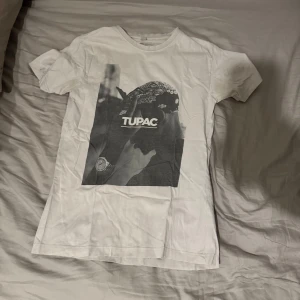 Vit T-shirt med Tupac-tryck - Snygg vit T-shirt med ett stort svartvitt tryck av Tupac på framsidan. Perfekt för dig som gillar klassisk hiphop-stil. T-shirten har en normal passform och korta ärmar.