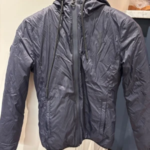 Svart vindjacka med huva - Snygg svart vindjacka med huva och dragkedja framtill. Perfekt för blåsiga dagar. Jackan har en stilren design med långa ärmar och är tillverkad i ett lätt material. Jackan är från cavalleria toscana, storlek 12, nypris 3000