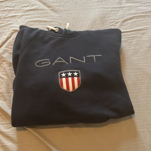 Mörkblå hoodie från GANT - Säljer en snygg mörkblå hoodie från GANT med broderad logga och emblem på bröstet. Tröjan har en praktisk känguruficka och dragsko i halsen. Perfekt för en avslappnad stil.
