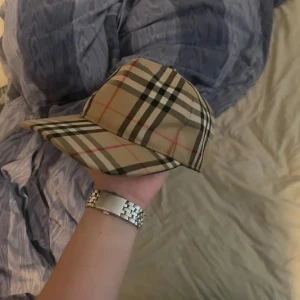  keps från Burberry - Snygg rutig keps från Burberry i beige, svart och röd. Klassisk design med justerbar rem baktill för perfekt passform. Perfekt för en stilren look.