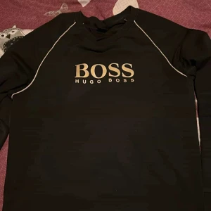 Svart långärmad tröja från Hugo Boss - Snygg svart långärmad tröja från Hugo Boss med guld logga på bröstet. Tröjan har guldiga detaljer längs ärmarna som ger en sportig look. Perfekt för en stilren och avslappnad stil. riktigt fet köpte här på plick för 1200 men passade inte mig tyvär, annars tyckte jag den va jävligt snygg