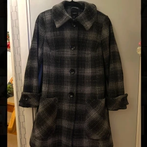 Rutig kappa från Lindex - Snygg rutig kappa från Lindex i svart och grått. Den har långa ärmar och stora knappar framtill. Perfekt för höst och vinter med sin klassiska stil och bekväma passform. Passar storlek M/L