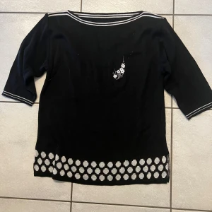 Svart tröja med blommönster - Snygg svart tröja med vita blommor längs nederkanten och en liten blomdetalj på bröstet. Tröjan har trekvartsärmar och vita ränder vid halsen och ärmsluten. Perfekt för en stilren look!