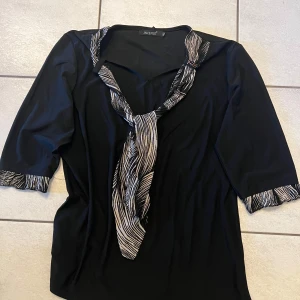 Svart blus med zebramönstrade detaljer från Orsay - Elegant svart blus från Orsay med zebramönstrade detaljer vid krage och ärmslut. Blusen har trekvartsärmar och en dekorativ knytning framtill. Perfekt för en stilren look.