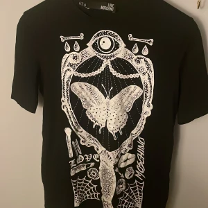 Svart t-shirt från Love Moschino - Cool svart t-shirt från Love Moschino med ett stort vitt tryck av en fjäril och olika symboler. T-shirten har korta ärmar och en normal passform. Perfekt för dig som vill sticka ut med en unik design som inte längre går att köpa.