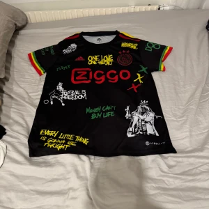 Svart fotbollströja Ajax med Bob Marley-tema - Cool svart fotbollströja från Adidas med Bob Marley-tema. Tröjan har färgglada detaljer i rött, gult och grönt på ärmarna och olika tryck med citat och bilder. Perfekt för fans av både fotboll och reggae. Pris kan diskuteras!