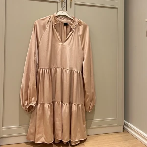 Champagnefärgad klänning från Gina Tricot - Elegant champagnefärgad klänning från Gina med v-ringning och volangdetaljer vid halsen. Klänningen har långa ärmar och en luftig passform. Använd en gång💘