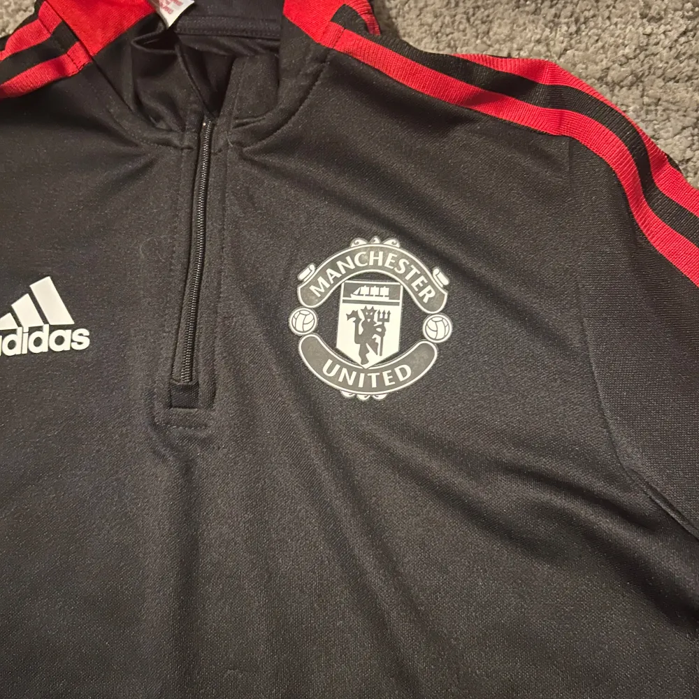 Säljer en svart långärmad Manchester United tröja från Adidas med röda detaljer och dragkedja vid halsen. Perfekt för fotbollsfans! Tröjan har klubbens emblem på bröstet och är tillverkad i Primegreen-material.. Neuletakit & Villapaidat.