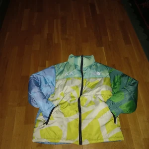 The north face puffer jacka som är flefärgad - Säljer en cool flerfärgad pufferjacka från The North Face. Jackan har ett unikt mönster i grönt, blått och gult med dragkedja framtill. Perfekt för kyliga dagar med sin quiltade design.