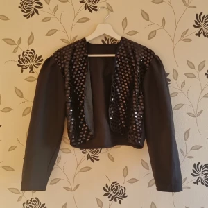 Svart bolero/överdel med paljetter, Strl: M/38 - Snygg svart bolero/överdel med glittrande paljetter på framsidan. Perfekt för att lägga till lite glamour till din outfit. Jackan har långa ärmar och är tillverkad i polyester. Tillverkad i Frankrike.