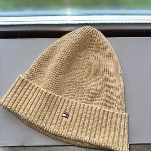 Beige stickad mössa från Tommy Hilfiger - Snygg beige stickad mössa från Tommy Hilfiger med ribbad kant. Perfekt för kyliga dagar och en stilren look. Den har en liten broderad logga framtill.