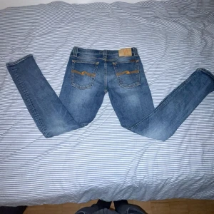 Blå jeans från Nudie Jeans - Snygga blå jeans från Nudie Jeans med klassisk femficksdesign och orange sömmar. De har en slim passform och är perfekta för en avslappnad stil. Tillverkade i slitstarkt denim. Modell tight long John!