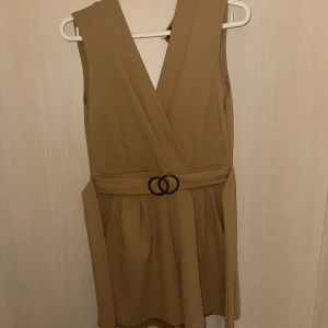 Beige ärmlös byxdress - Snygg beige byxdress med ett elegant bälte i midjan och en stilren rosett bak. Byxdressen är ärmlös och har en klassisk omlottdesign som ger en smickrande passform.