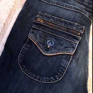 Mörkblå jeans med broderad ficka - Snygga mörkblå jeans med unik broderad ficka och dragkedja. Perfekta för en cool och avslappnad stil. Jeansen har en klassisk passform och är tillverkade i slitstarkt denim.