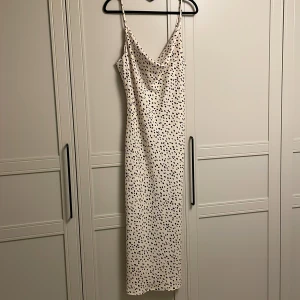 Prickig långklänning med smala axelband - Snygg långklänning i beige med svarta prickar. Klänningen har smala axelband och en v-ringad design som ger en elegant look. Perfekt för festliga tillfällen. 131 cm lång från axelbanden och ner, axelbanden går att justera med. Är oanvänd. 