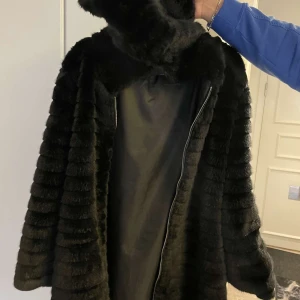 Svart pälsjacka från J&J - Snygg svart pälsjacka från J&J med dragkedja framtill. Jackan har en elegant design med långa ärmar och är perfekt för kyligare dagar. Den mjuka pälsen ger en lyxig känsla och stil.