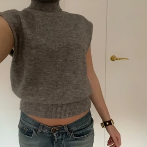 Grå ärmlös polotröja - Säljer en stilren grå ärmlös polotröja i mjukt material. Tröjan har en ribbad kant vid midjan och en hög polokrage. Köpt vintage. Endast använd ca 2gånger 