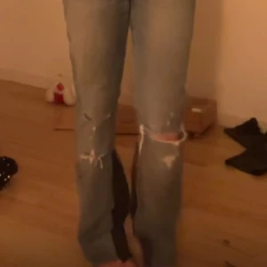 Blå jeans med slitningar - Säljer ett par blå jeans med slitningar på knäna och en unik mörkare lapp på bakfickan. De har en klassisk passform och är perfekta för en avslappnad stil.