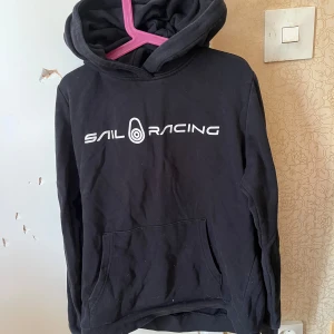 Svart hoodie från Sail Racing - Säljer en svart hoodie från Sail Racing i storlek 160. Den är superbekväm och perfekt för både vardag och chill. Hoodien har en stor ficka fram och en klassisk logga i vitt på bröstet. Perfekt för höst och vinter!
