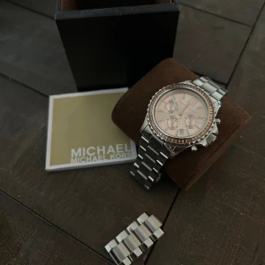 Silverfärgad klocka från Michael Kors - Säljer en snygg och stilren klocka från Michael Kors. Den har en silverfärgad länkarmband och en urtavla med romerska siffror och tre mindre urtavlor för extra funktioner. Klockan har en glittrig kant runt urtavlan som ger den en extra touch av elegans. champange färgade bakgrund. Kommer med en extra länk. Perfekt för både vardag och fest!