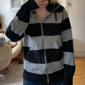 Randig hoodie med dragkedja - Säljer en mysig randig hoodie i svart och grått. Den har en dragkedja framtill och en justerbar huva med snören. Tröjan är i välanvänt skick med några tecken på användning, men fortfarande superbekväm.