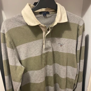 GANT Heavy Rugbytröja   - Säljer en stilren och robust GANT heavy rugbytröja i storlek S. Tröjan är grön och grå randig med en kontrasterande off-white krage. Tröjan är i mycket gott skick och visar inga tecken på slitage. Nypriset för denna tröja var 1500 SEK.