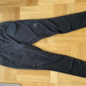 Svarta träningsbyxor från Adidas - Säljer ett par svarta träningsbyxor från Adidas. De är i bra skick och perfekta för träning eller en avslappnad dag. Byxorna har en smal passform och är gjorda i ett bekvämt syntetmaterial. Adidas-loggan syns på sidan.