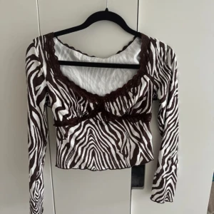 Zebramönstrad topp med spetsdetaljer - Säljer en supercool zebramönstrad topp med långa ärmar. Toppen har snygga bruna spetsdetaljer runt halsen och under bysten, vilket ger den en unik look. Perfekt för en utekväll eller fest!