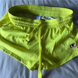 Gula shorts från Champion - Säljer ett par coola gula shorts från Champion. De är i storlek medium och har en bekväm elastisk midja med knytband. Perfekta för sommaren och träning!