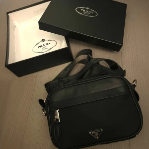 Prada väska - Säljer en snygg svart axelväska från Prada. Väskan är i nyskick och kommer med originalförpackning. Den har en justerbar axelrem med Prada-loggan och en praktisk dragkedja framtill. Perfekt för både vardag och fest! 