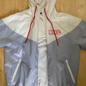 Grå och vit vindjacka från Nike - Säljer en snygg grå och vit vindjacka från Nike. Jackan har en cool design med röda detaljer och dragkedjor på fickorna. Perfekt för vår och höst när det blåser lite extra. Den har en huva med röda snören för att justera passformen.