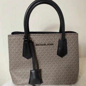 Beige och svart handväska från Michael Kors - Säljer en snygg och stilren handväska från Michael Kors. Väskan är beige med ett diskret MK-mönster och har svarta detaljer. Den har två rejäla handtag och en praktisk dragkedja inuti för säker förvaring. Perfekt för både vardag och fest! fler bilder kan skickas, liten mindre modell