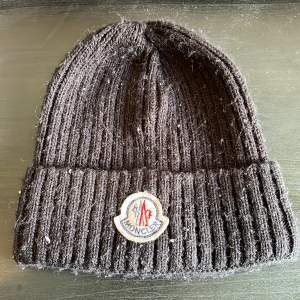 Säljer en svart ribbad mössa från Moncler. Den är gjord av 100% bomull och har en broderad Moncler-logga framtill. Mössan är perfekt för kyliga dagar och har en klassisk stil som passar till det mesta. Den är i välanvänt skick men fortfarande supermysig!