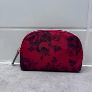 Röd blommig necessär från Victoria's Secret - Säljer en superfin röd necessär från Victoria's Secret med ett vackert blommönster. Den har en praktisk dragkedja och är perfekt för att förvara smink eller andra småsaker. Insidan är randig och ger en lyxig känsla. Perfekt för resor eller som en söt accessoar i badrummet eller i din axelarm väska 