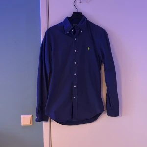 Blå skjorta från Polo Ralph Lauren - Snygg blå skjorta från Polo Ralph Lauren med en broderad grön logga på bröstet. Skjortan har en slim passform och knappar framtill. Perfekt för en stilren look.