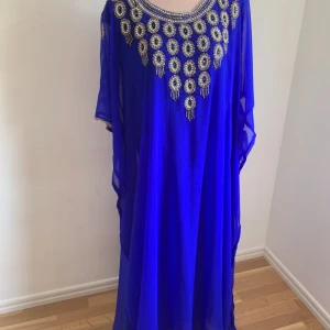 Blå kaftan med dekorativa detaljer - Elegant blå kaftan med vackra dekorativa detaljer runt halsringningen. Klänningen har en lös passform och är tillverkad i ett lätt och flödande material. Perfekt för en speciell kväll eller festlig tillställning.