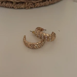 Guldiga örhängen med glittriga detaljer - Snygga guldiga örhängen i creol-stil med glittriga stenar som ger en lyxig känsla. Perfekta för att lyfta vilken outfit som helst med lite extra bling. De har en elegant vriden design som fångar ljuset fint.