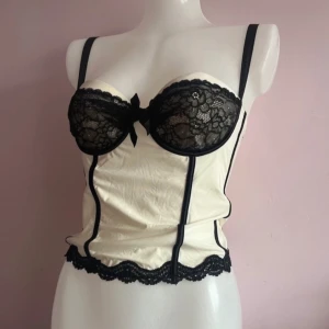 Vit korsett med svarta spetsdetaljer - supercool vit korsett/topp med svarta spetsdetaljer och rosett framtill. den har justerbara axelband samt vid ryggen 💓85b kupa!!