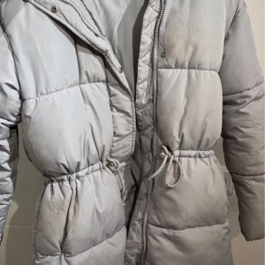 Grå dunjacka från H&M - Säljer en stilren grå dunjacka från H&M i storlek XS. Jackan har en dragkedja framtill och justerbar midja med knytband för en bättre passform. Perfekt för kyliga dagar med sin långa ärm och vadderade design.
