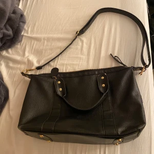 Svart axelväska med dragkedja - Snygg svart axelväska i skinn med praktisk dragkedja och justerbar axelrem. Väskan har även handtag och en yttre ficka med dragkedja. Insidan är rymlig med röd foder, perfekt för att organisera dina saker.  Endast ett få antal gånger, så den är fortfarande i ett bra skick. Säljer då den inte kommer till användning längre. Har även 3st små innefack och 1st på baksidan av väskan. OBS! Jag har även lagt ut annonsen på Vinted också