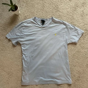 Ljusblå t-shirt från Polo Ralph Lauren - Säljer en ljusblå t-shirt från Polo Ralph Lauren med en liten gul logga på bröstet. T-shirten är tillverkad i 100% bomull och har en klassisk rund halsringning. Perfekt för en avslappnad stil.