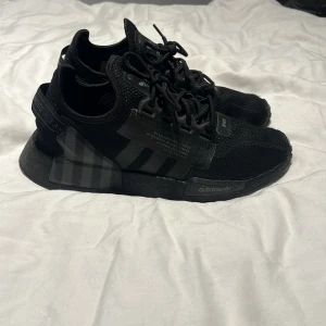 Svarta Adidas NMD sneakers - Säljer ett par svarta Adidas NMD sneakers med en stilren design. Skorna har en stickad ovandel och klassiska tre ränder på sidan. De är utrustade med snörning och har en bekväm sula för optimal komfort.