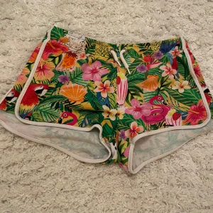Färgglada blommiga shorts från H&M - Säljer ett par färgglada shorts från H&M i samarbete med Emma Jayne. De har ett tropiskt mönster med blommor, flamingos och papegojor. Shortsen har vita kanter och en snörning i midjan. Funkar som shorts o vardagen eller som pyjamas shorts😊 Använd ett fåtal gånger men blev snabbt urvuxna tyvärr🩷 Köpare står för frakt!