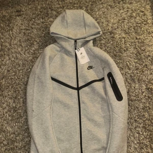 Grå hoodie från Nike - Säljer en helt ny oanvänd grå hoodie från Nike med dragkedja och huva. Den har en svart Nike-logga på bröstet och en praktisk ficka med dragkedja på ärmen. Perfekt för en sportig look!