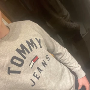 Grå sweatshirt från Tommy Jeans - Säljer en stilren grå sweatshirt från Tommy Jeans med deras ikoniska logga i svart, vitt och rött på bröstet. Tröjan har långa ärmar och ribbade muddar vid ärmslut och nederkant. Perfekt för en avslappnad look. Skriv gärna vid minsta fundering!