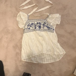 Vit blus med blå broderier - Charmig vit blus med blå broderier och korta puffärmar. Blusen har en fyrkantig halsringning och knytband vid axlarna. Perfekt för en somrig look!