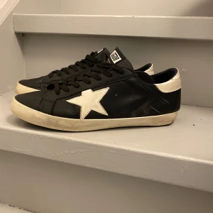Svarta sneakers med vit stjärna - Snygga svarta sneakers från GGDB med en vit stjärna på sidan. Skorna har svarta snören och en vit sula som ger en cool kontrast. Perfekta för en avslappnad stil.Skorna är i ett helt okej skick. Har haft dem i snart 2 år så dem är slitna nu vi har defekter, de är även därför priset är lågt. Saknar kvitto men allt annat og medföljer 