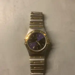 Snygg Omega klocka med en unik blå/lila urtavla och gulddetaljer. Armbandet är i silver och guld, vilket ger en elegant touch. Perfekt för den som vill ha något stilrent och klassiskt. Säljer den billigt då jag inte använder den och vill sälja snabbt.